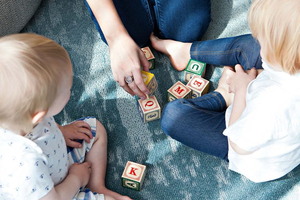 The image shows a group of people playing with cards. There is a person wearing clothing and a toddler involved in the activity. A baby, a boy, and a child are also present in the scene, they could be autistic or not. The setting appears to be indoors and conducting therapies.