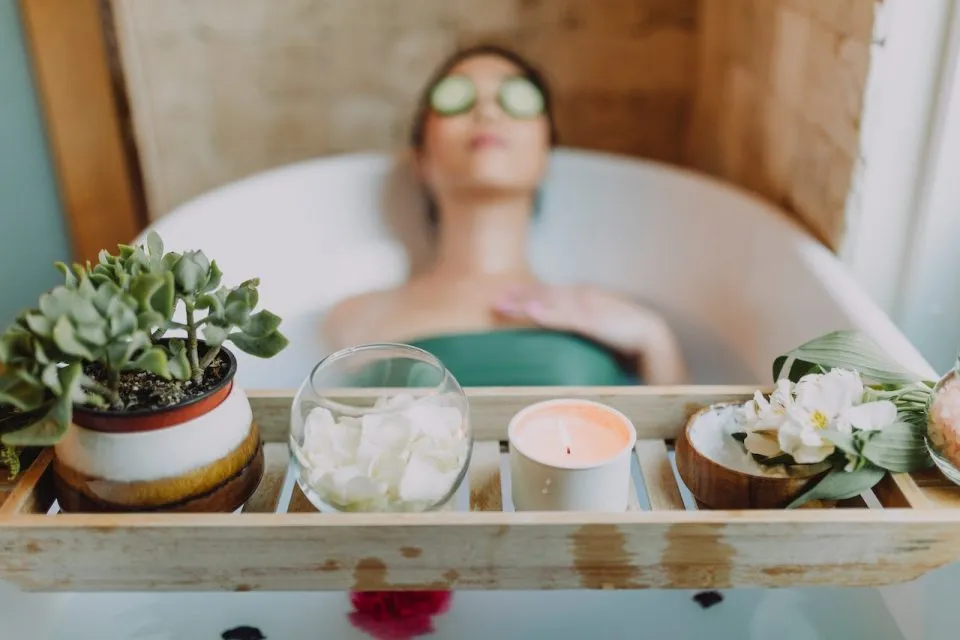 Someone laying back in a bath with a plant, candle, and bowl on a bath caddy in the foreground.