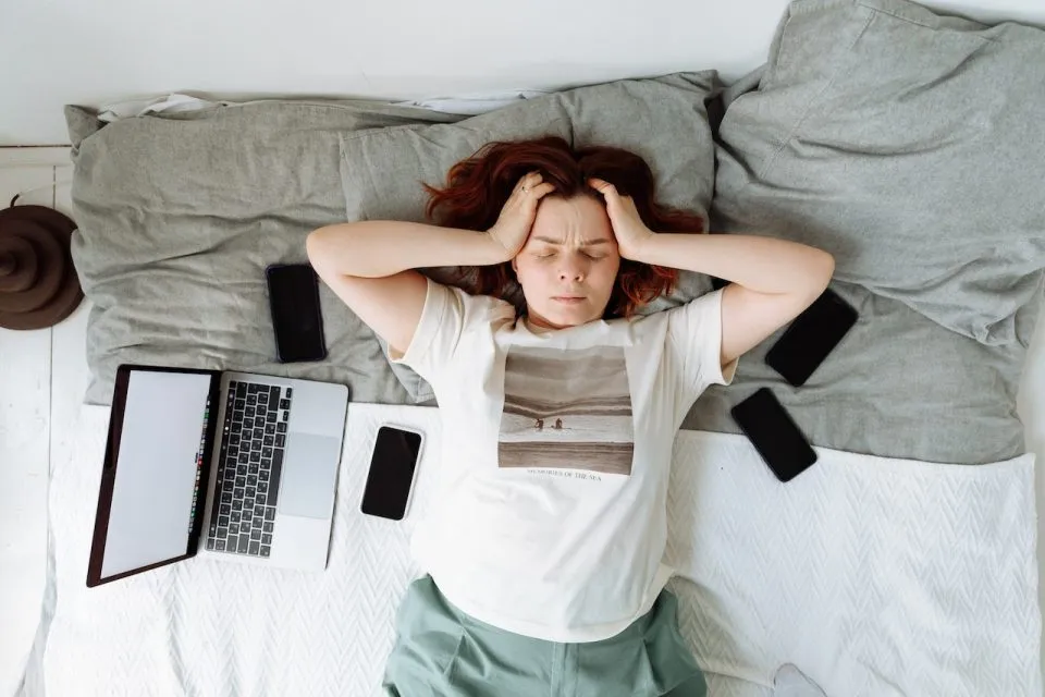 A person looking stressed out in bed surrounded by phones and a laptop. Her hands are on her head and her eyes are closed. 