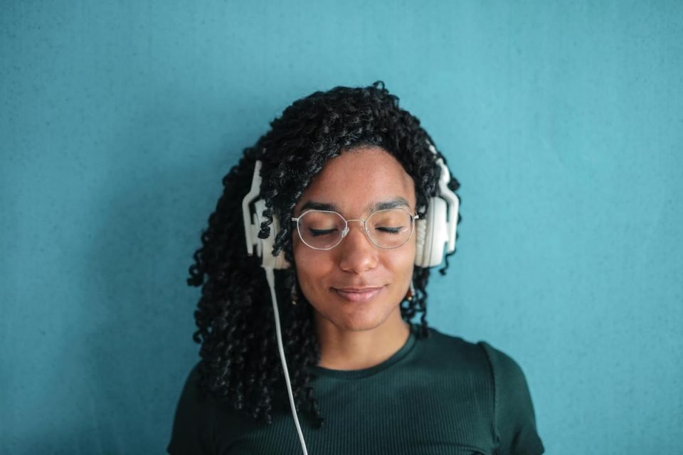 A person closing their eyes with over-ear headphones on. They're in front of a blue background wearing a dark blue shirt. (Andrea Piacquadio, Pexels)