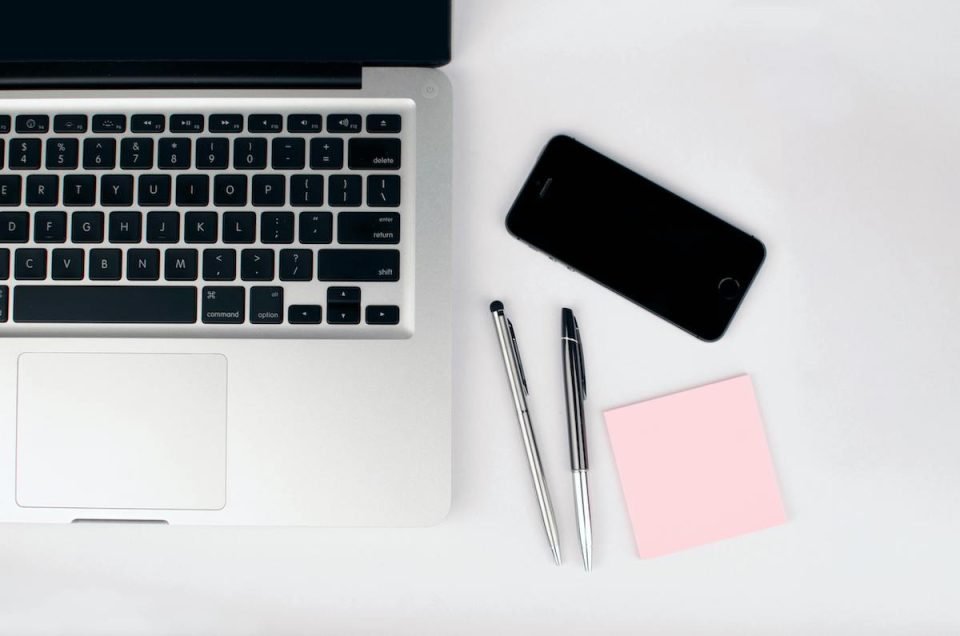 A Macbook on a desk next to pens, a cell phone, and a memo pad. (Lukas/Pexels)