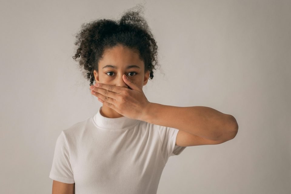 A person shown from the waist up covers their mouth with one hand. They're wearing a white shirt and standing against a beige background.