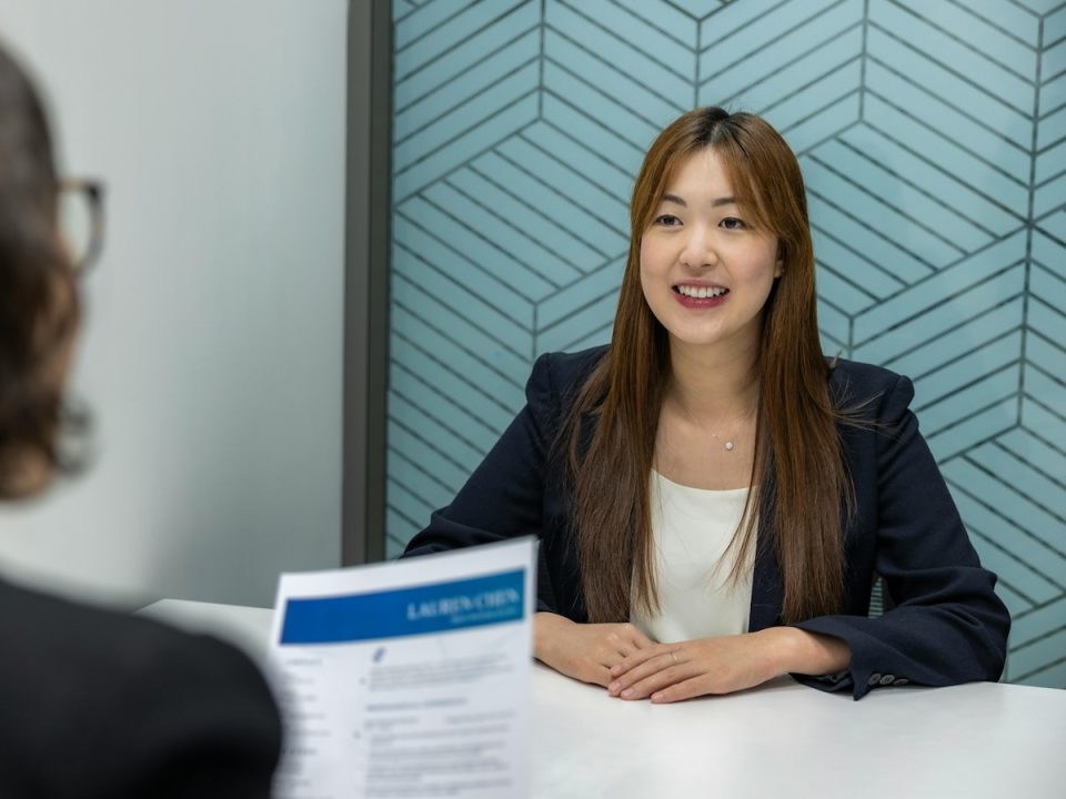 A person sits across from an interviewer at a table. They are on a blue background and wearing a dark blazer and white shirt.
