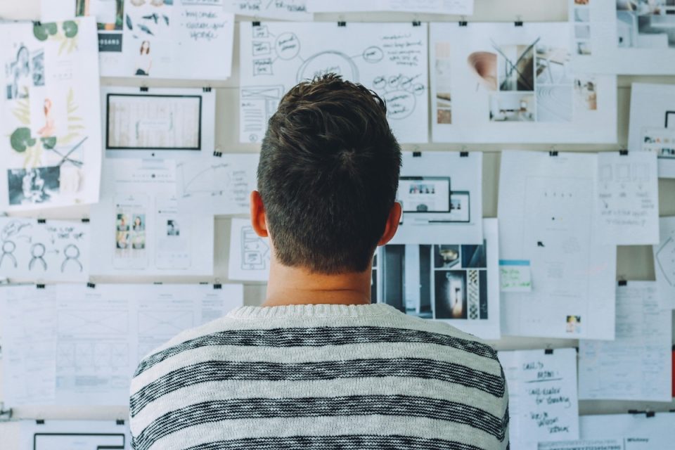 Someone looking at a wall full of research. They're facing away from the camera with a short haircut wearing a striped shirt. (Pexels/Startup Stock Photos)