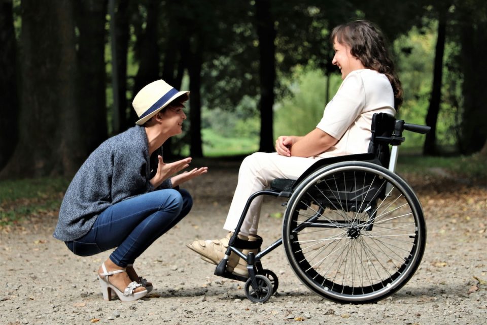 One person kneels to talk face-to-face with another person in a wheelchair. They're outside and surrounded by trees on a trail. (Judita Mikalkevičė/Pexels)