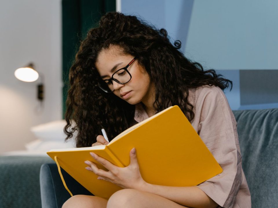 A woman journalling in her home. She's wearing glasses and wearing a beige shawl, and the journal is bright yellow. (Canva)