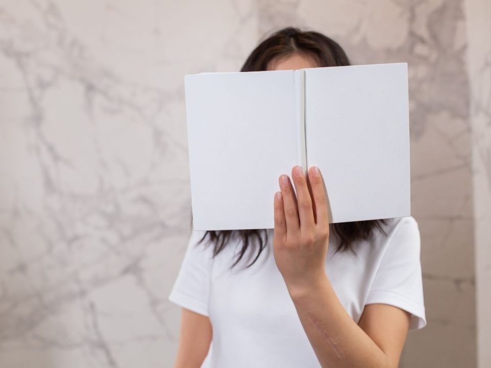 A woman hiding her face with a book. Her shirt and the book cover are both all white. (Canva)