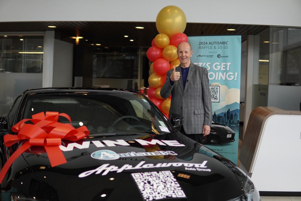 A man gives a thumbs-up beside a black car with a large red bow. Colorful balloons and a raffle poster are in the background, creating a celebratory atmosphere.
