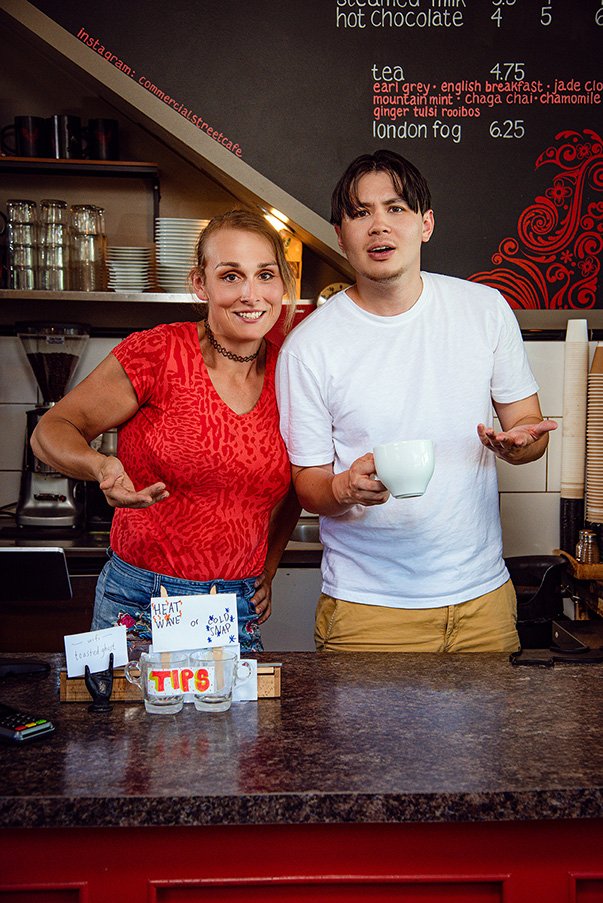 A woman in a red shirt and a man in a white shirt stand smiling behind a cafe counter. He holds a mug, with menu prices visible on the wall. The scene is friendly and inviting.