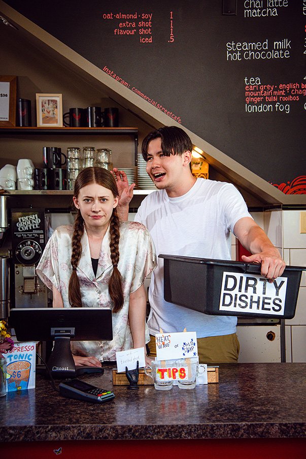 A woman with braided hair looks uncomfortable behind a café counter, while a cheerful man holds a box labeled "Dirty Dishes." Menu items are visible on a chalkboard behind them.