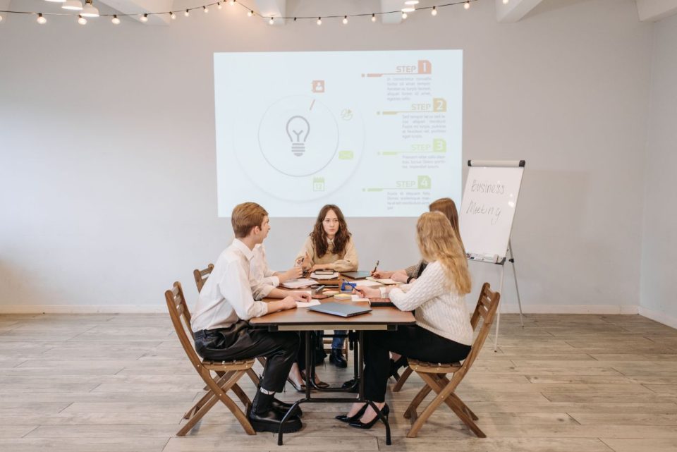 Young adults in a business meeting with infographics projected on a screen behind them. (Pavel Danilyuk/Pexels)