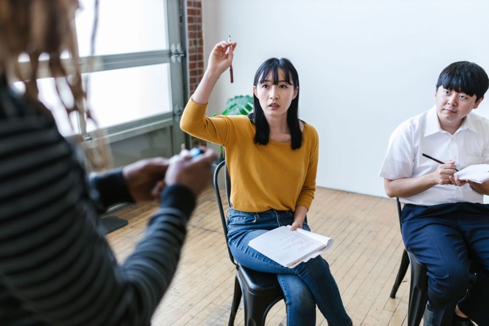A woman raising her hand to ask a question during a business meeting. She's holding a notebook and looking curiously and tentatively at a speaker out of frame. (RDNE Stock Project/Pexels)